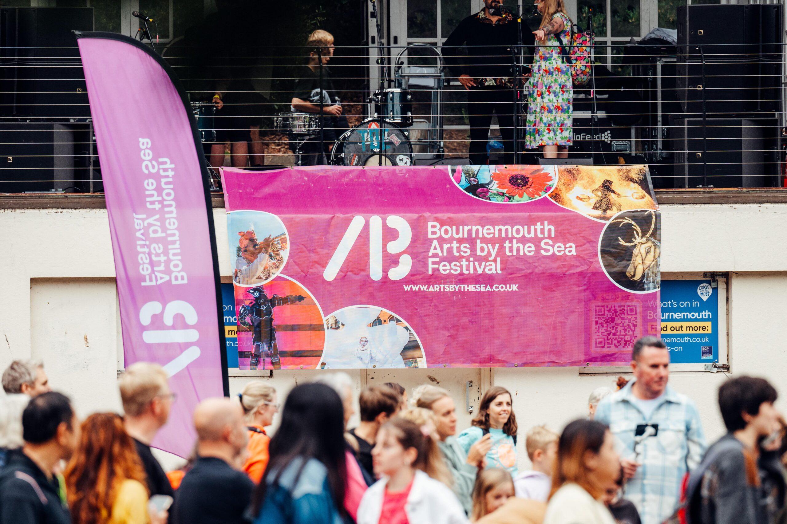A vibrant scene at the Bournemouth Arts by the Sea Festival, with a large pink banner prominently displayed. The banner features the festival logo, website, and colorful images of performers and artwork. In front of the banner, a crowd of people is gathered, with some blurred in motion. To the left, a purple flag with the festival's branding is visible, while a stage with musicians and performers can be seen in the background.