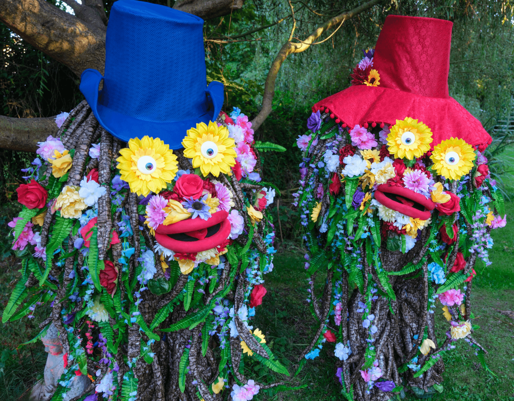 Two whimsical tree-like costumes decorated with colorful flowers and vines stand outdoors on grass. Each has exaggerated red lips, large googly eyes made of yellow flowers, and wears a tall hat—one blue, one red. The scene is playful and vibrant, set beneath leafy trees.