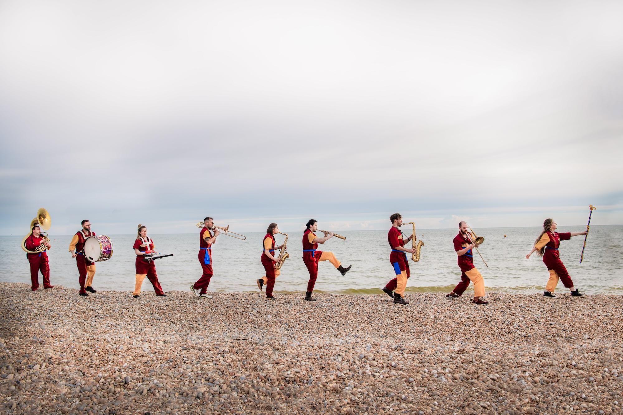 Brass band in colorful uniforms marching along a pebbled beach by the sea.