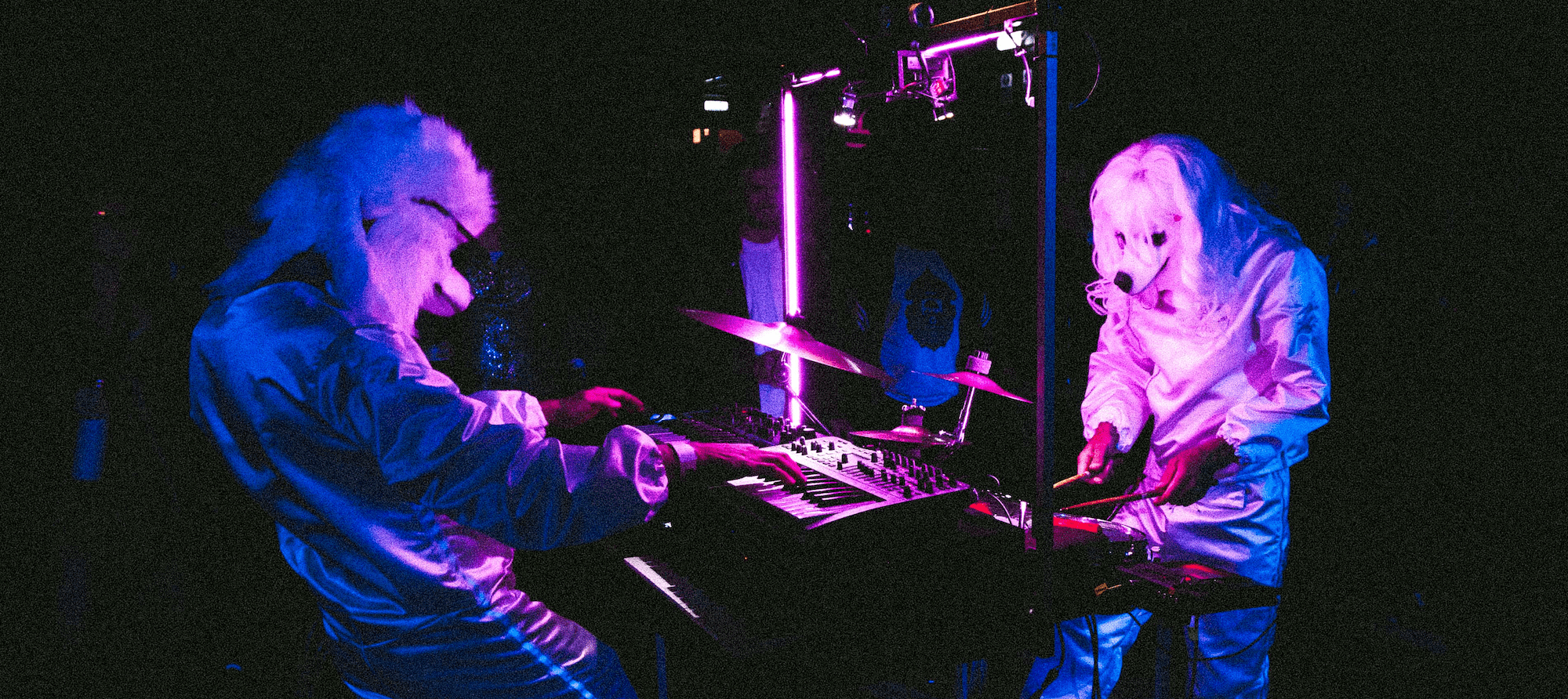 Two performers in white dog costumes play electronic instruments under neon lights.