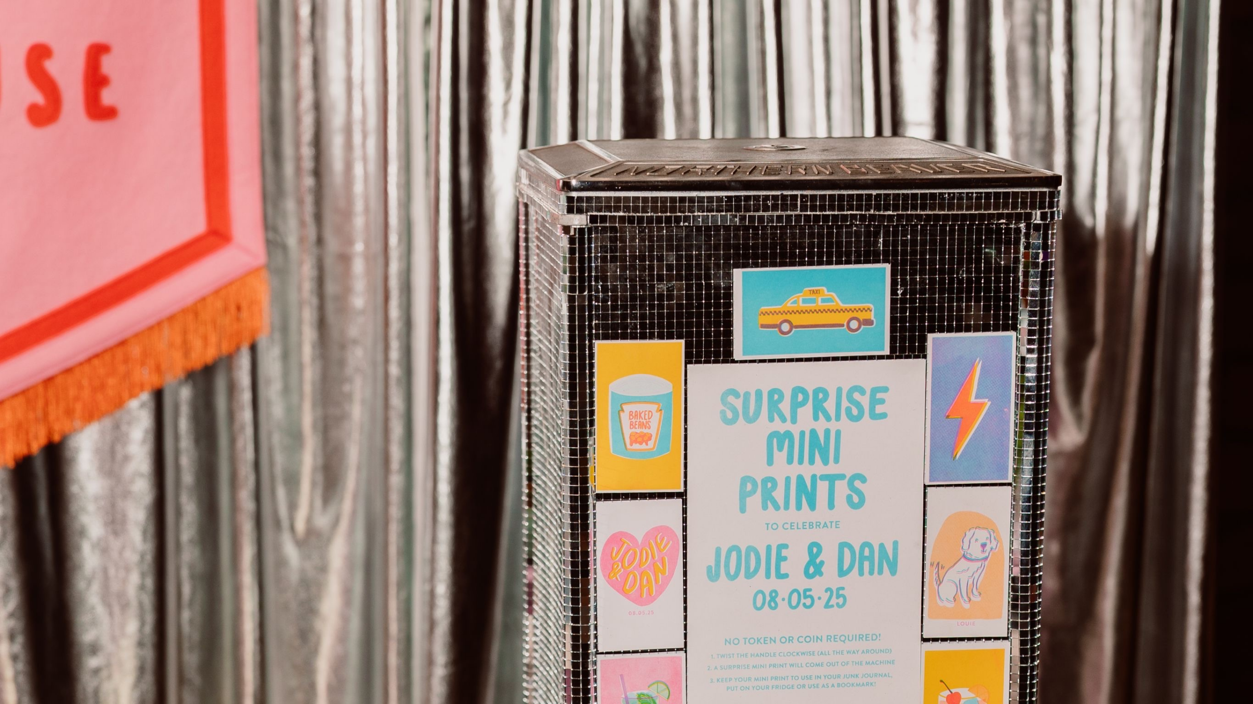 A shiny silver vending-style machine decorated with colorful stickers stands against a metallic curtain backdrop.