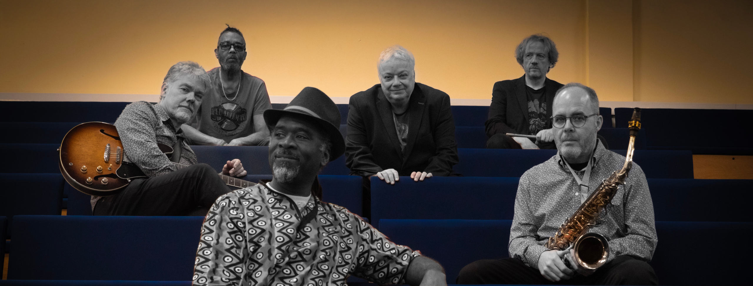 Six musicians sit in blue auditorium seats with guitar and saxophone, shown in a stylized photo with selective color highlights.