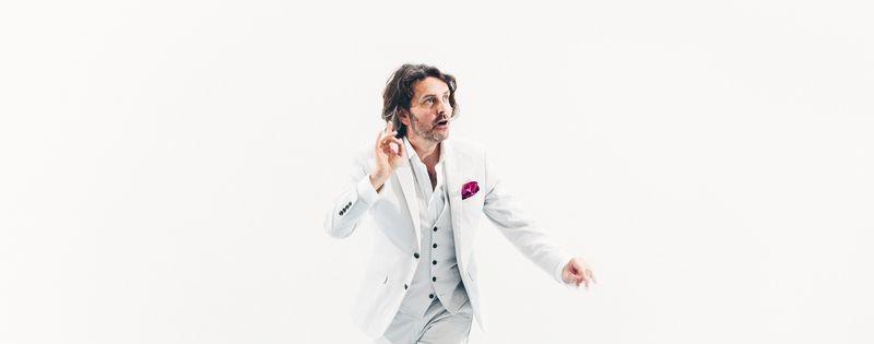 Man in a white suit with magenta pocket square gestures expressively against a plain white background, as if mid-performance.
