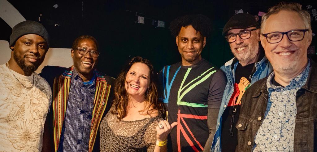 Six smiling people pose together in colorful, casual outfits, standing closely as a cheerful group portrait.