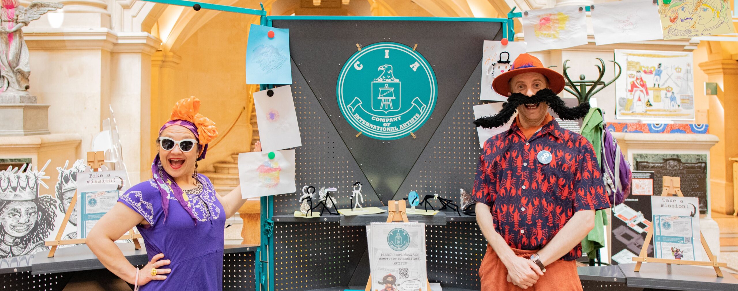 Two performers in bright costumes stand by a “Company of International Artists” display decorated with drawings and signs.
